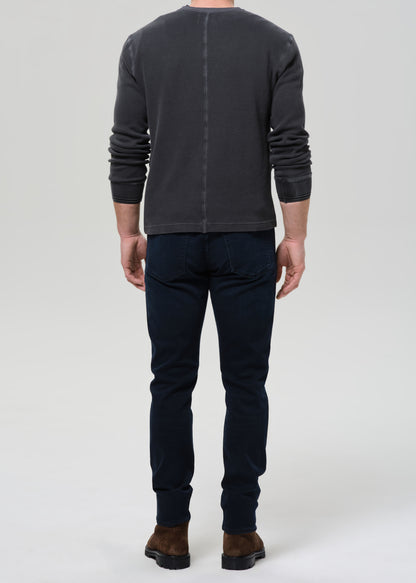 A man viewed from the back wearing a dark charcoal grey  long-sleeve crewneck shirt, dark navy blue jeans, and brown boots, standing against a grey  background.
