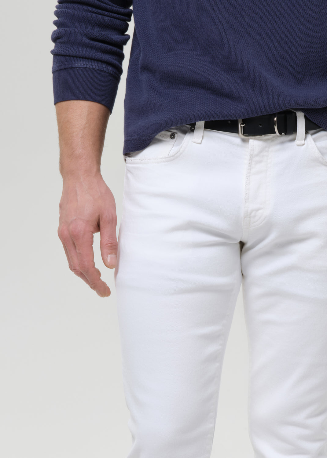 A close-up of a person wearing white slim-fit jeans, a black belt, and a navy blue ribbed long-sleeve top against a grey background.