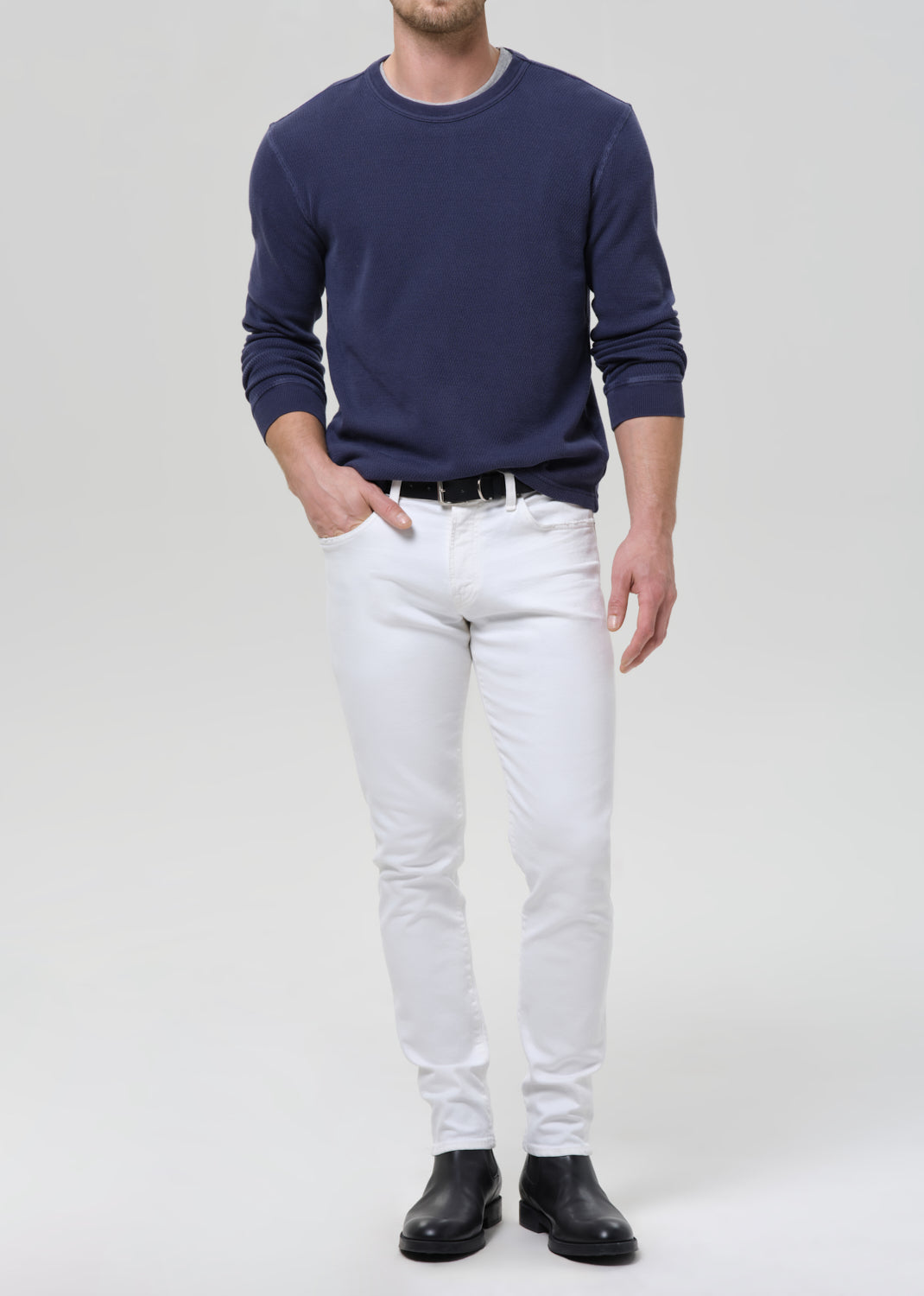 A man wearing a navy blue long-sleeve crewneck sweater, white slim-fit jeans, a black belt, and black leather boots, standing against a grey background.