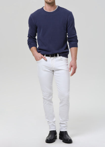 A man wearing a navy blue long-sleeve crewneck sweater, white slim-fit jeans, a black belt, and black leather boots, standing against a grey background.