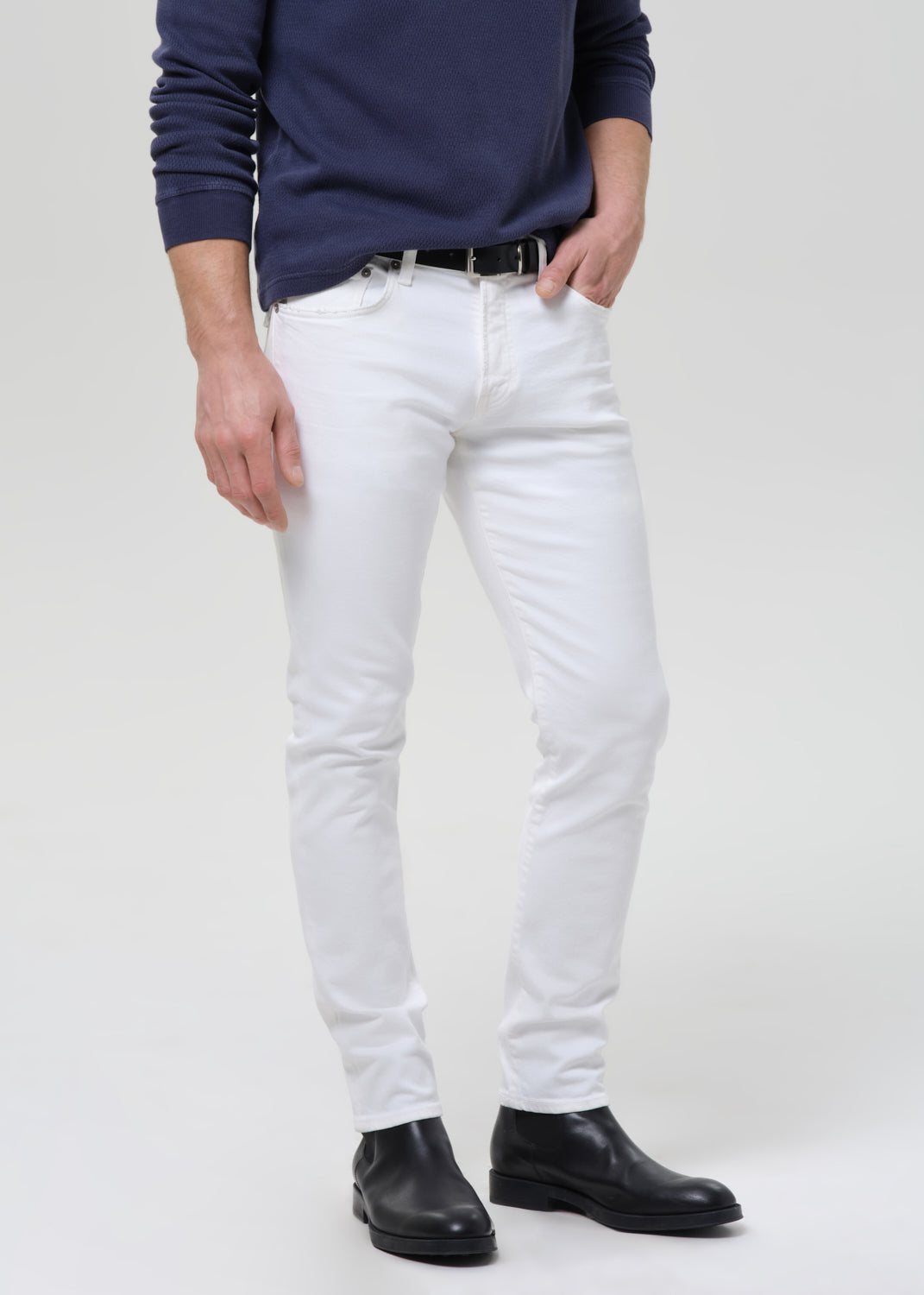 A man wearing slim-fit white jeans, a navy blue long-sleeve shirt, a black belt, and black leather boots, standing against a grey background.