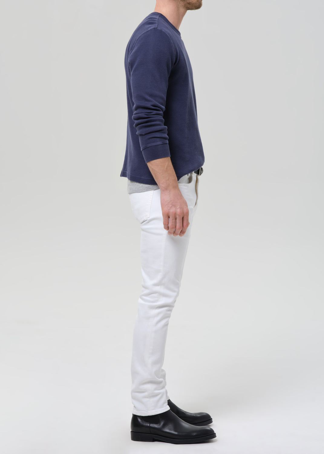 A man shown in profile wearing a navy blue long-sleeve shirt, white trousers, and black leather shoes against a grey background.