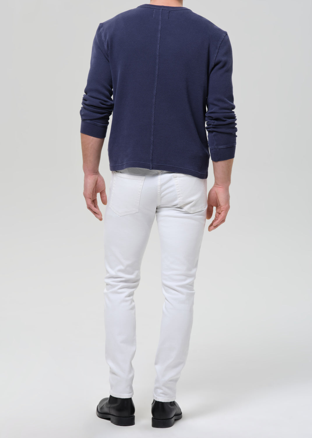 A man viewed from behind wearing a navy blue long-sleeve shirt, white slim-fit jeans, and black shoes, standing against a grey background.