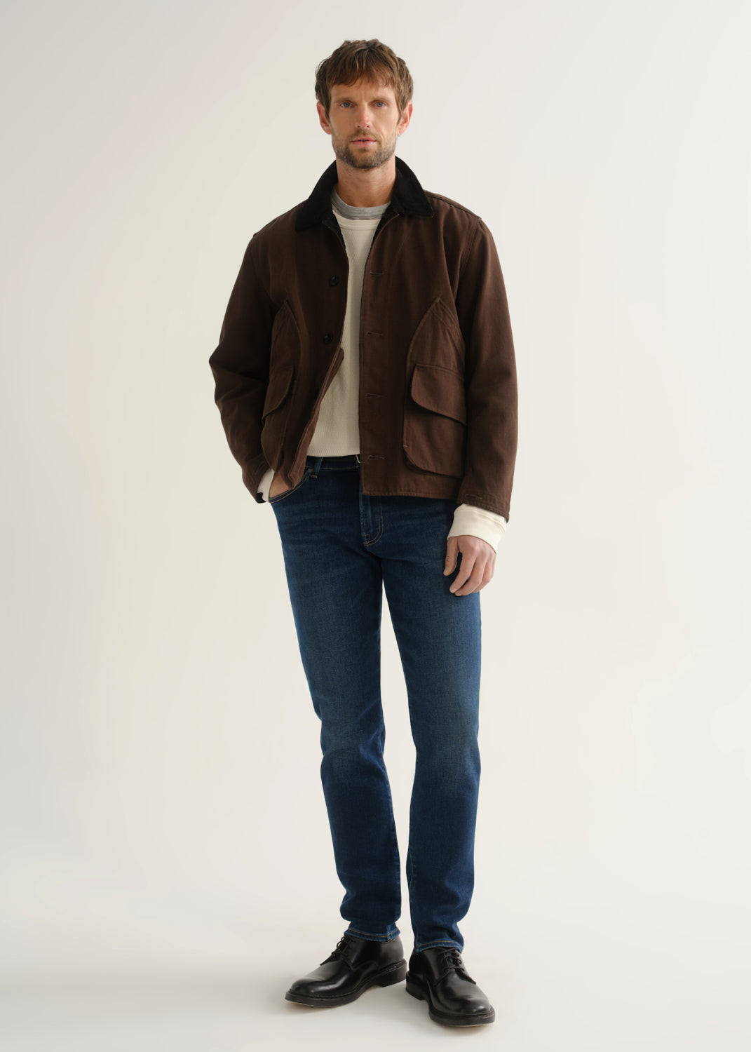 Person wearing medium blue slim tapered jeans with a brown jacket, cream sweater, and black shoes, front view.