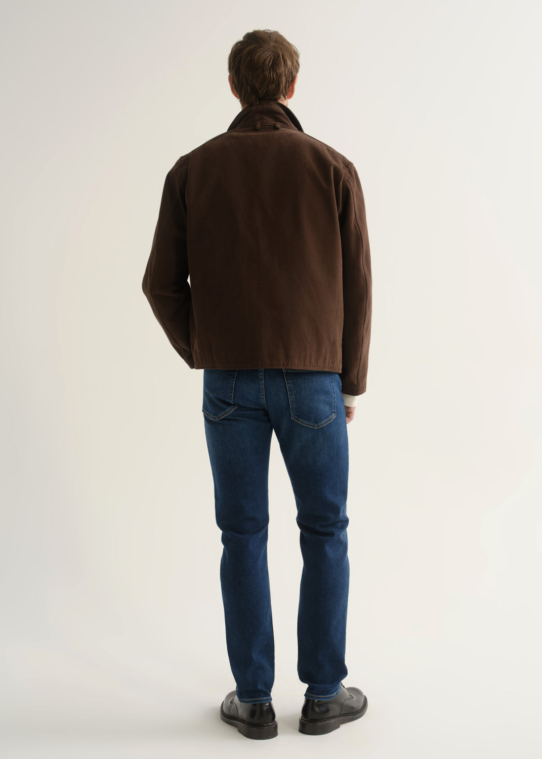 Person wearing medium blue slim tapered jeans with a brown jacket, cream sweater, and black shoes, front view.