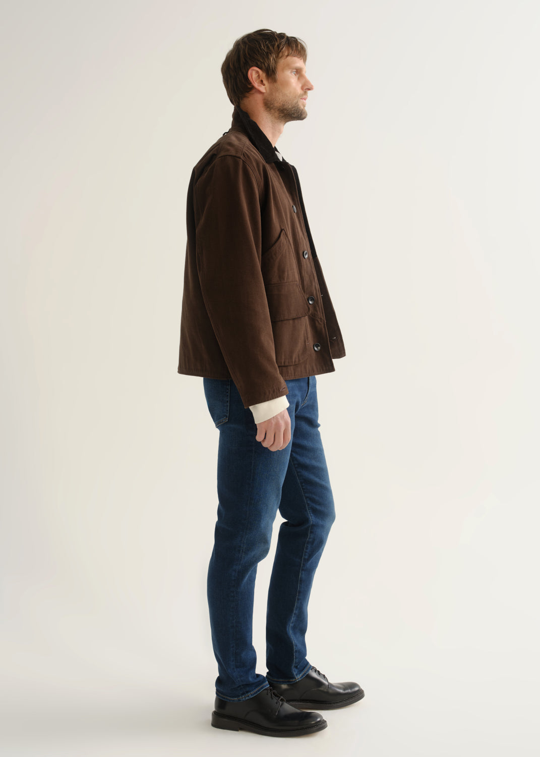 Person wearing medium blue slim tapered jeans with a brown jacket, cream sweater, and black shoes, side view.