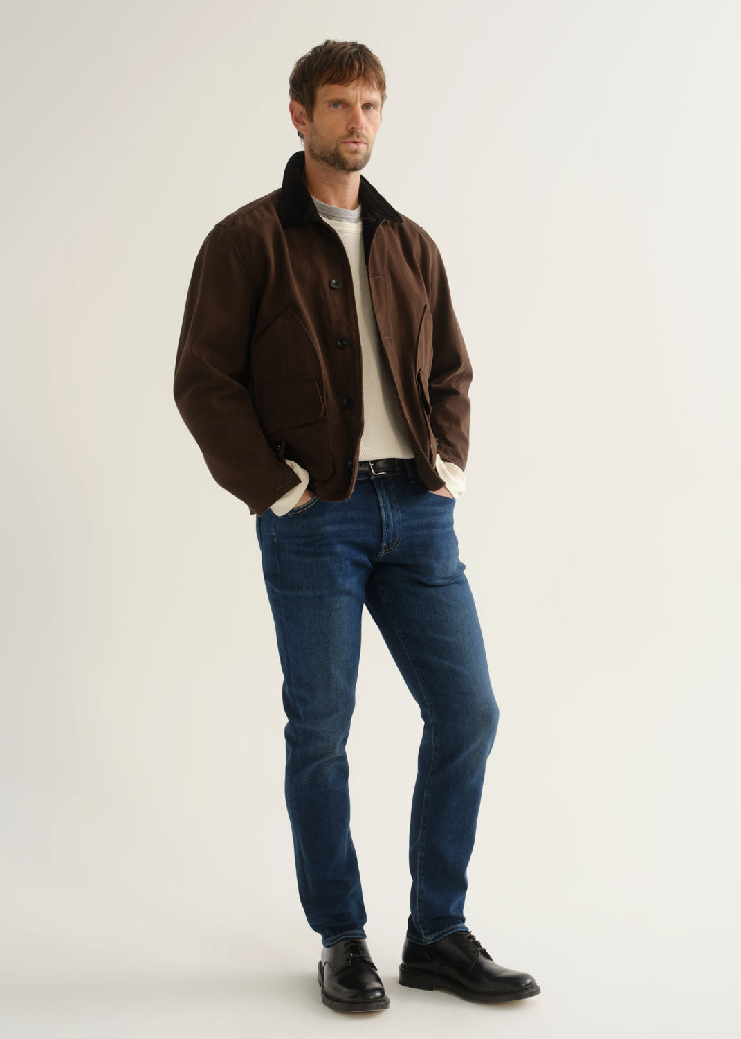Person wearing medium blue slim tapered jeans with a brown jacket, cream sweater, and black shoes, front angled view.