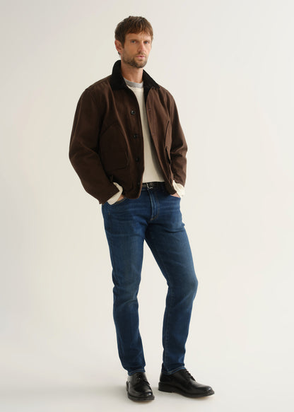 Person wearing medium blue slim tapered jeans with a brown jacket, cream sweater, and black shoes, front angled view.
