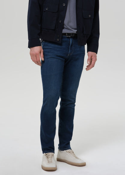 Man wearing medium blue straight leg jeans with a navy jacket and grey crewneck tee on a grey background.