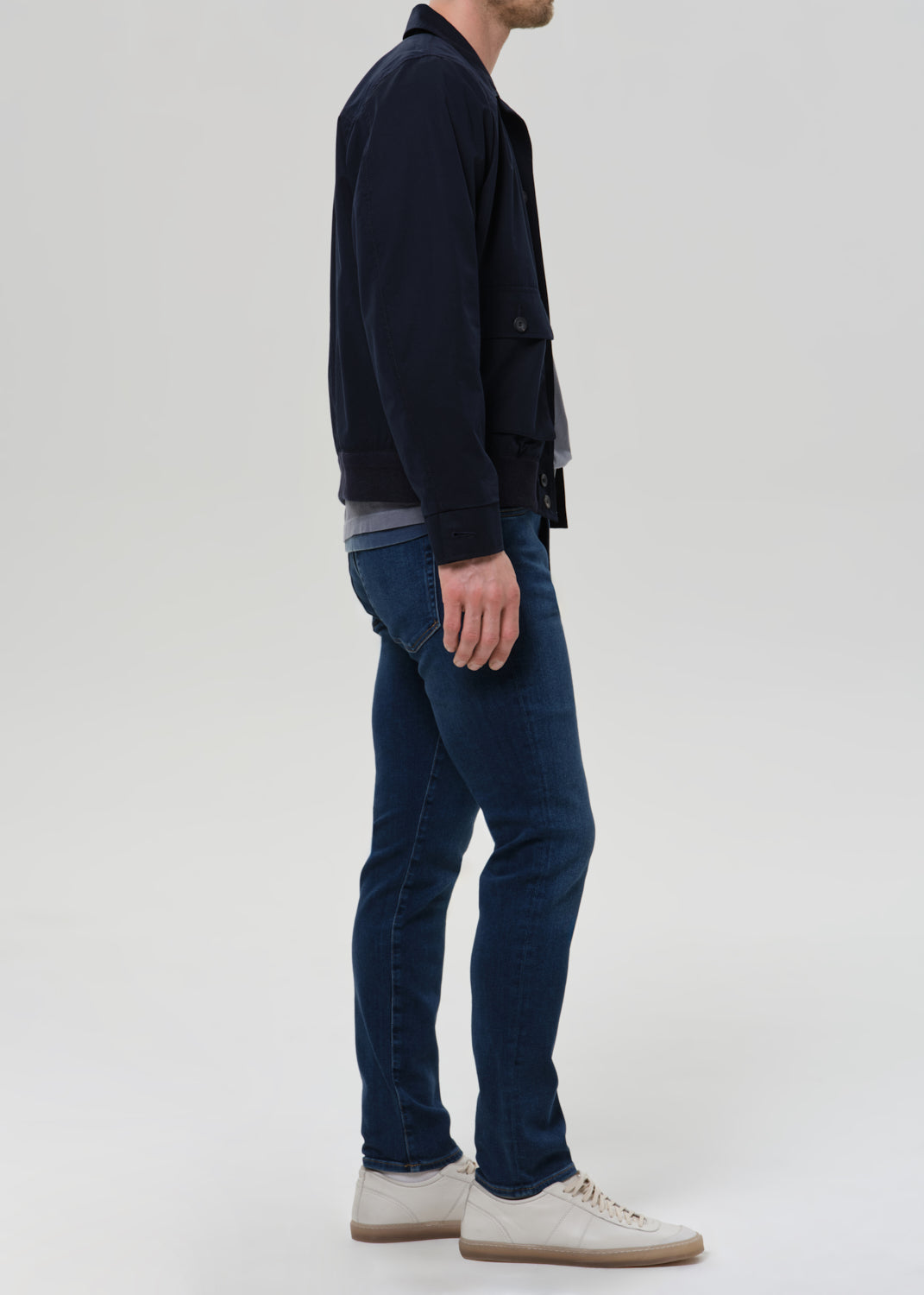 Man wearing medium blue straight leg jeans with a navy jacket and grey crewneck tee viewed from the side on a grey background.