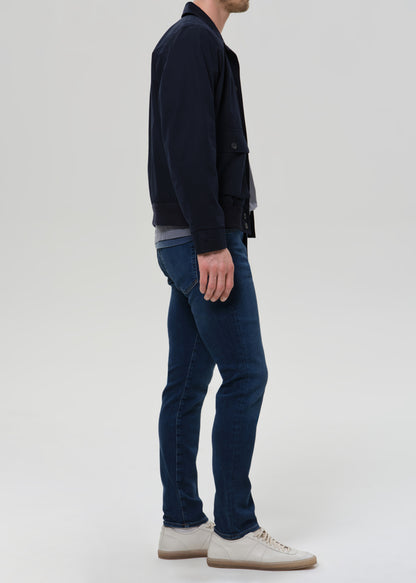 Man wearing medium blue straight leg jeans with a navy jacket and grey crewneck tee viewed from the side on a grey background.