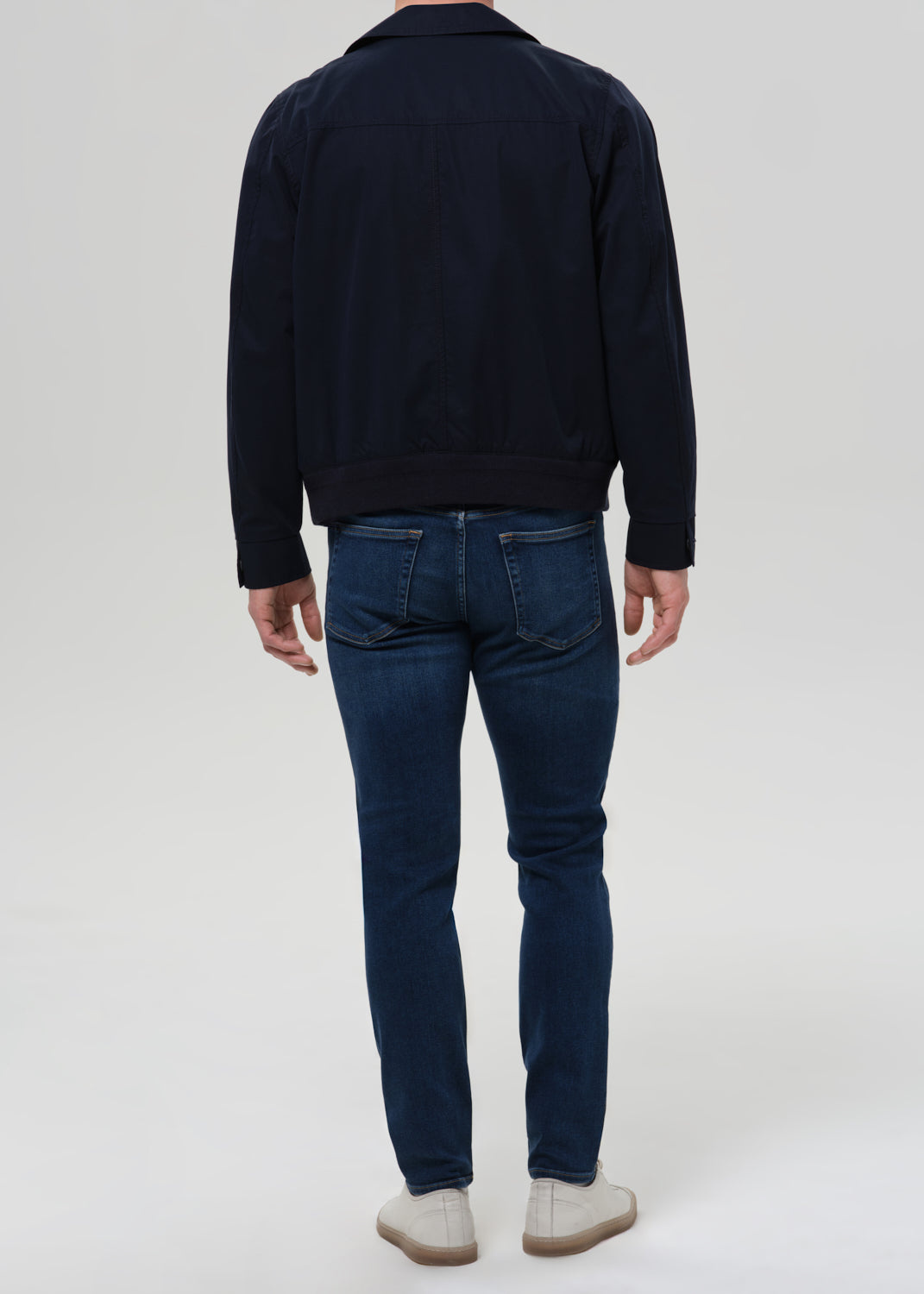 Man wearing medium blue straight leg jeans with a navy jacket and grey crewneck tee viewed from the back on a grey background.