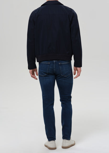 Man wearing medium blue straight leg jeans with a navy jacket and grey crewneck tee viewed from the back on a grey background.