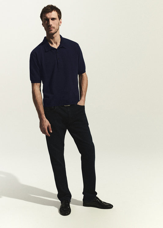 Man wearing a dark blue polo shirt and dark wash jeans on a white background