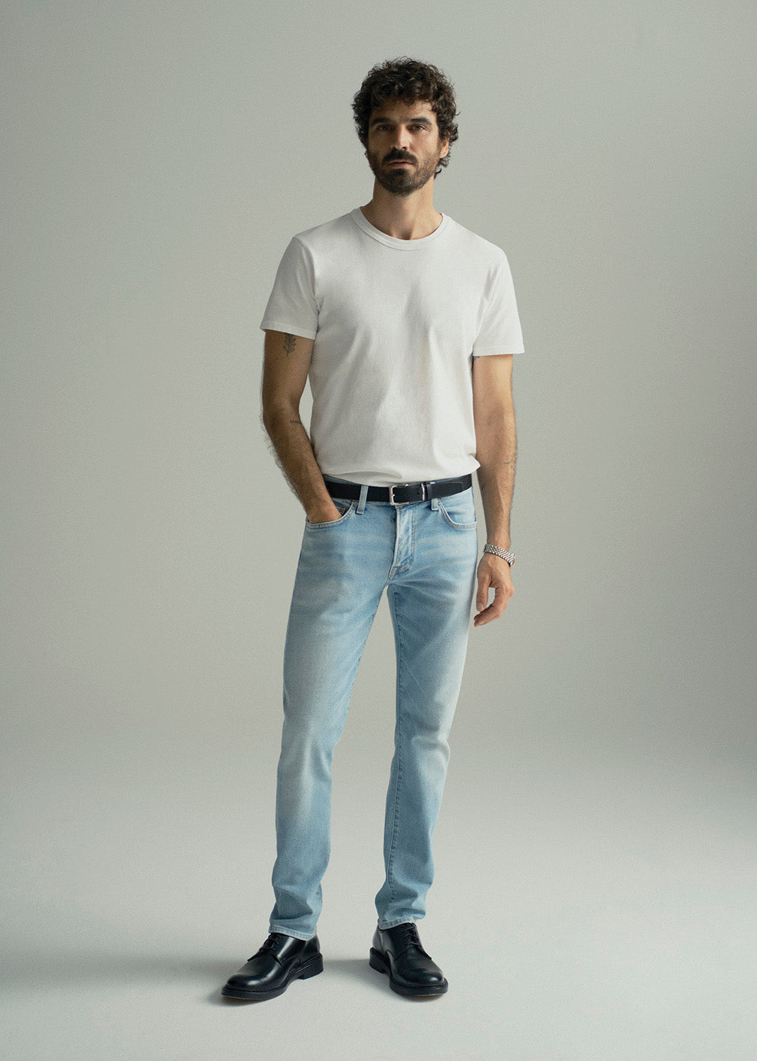 A man modeling a white crew-neck t-shirt, light-wash blue jeans, a black belt, and black leather shoes against a neutral gray studio background.