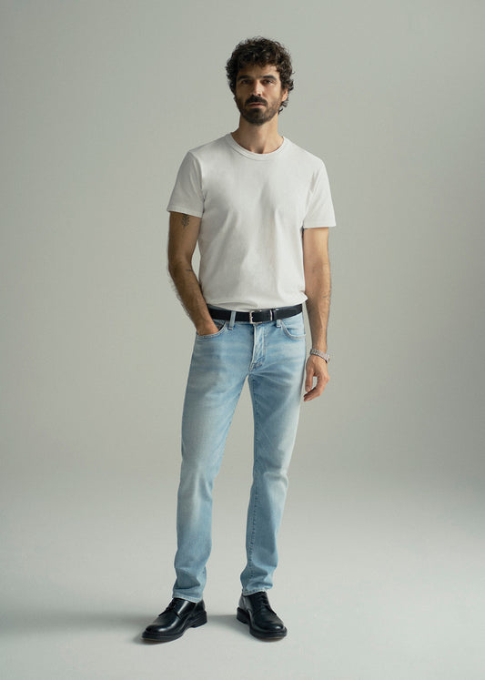 A man modeling a white crew-neck t-shirt, light-wash blue jeans, a black belt, and black leather shoes against a neutral gray studio background.