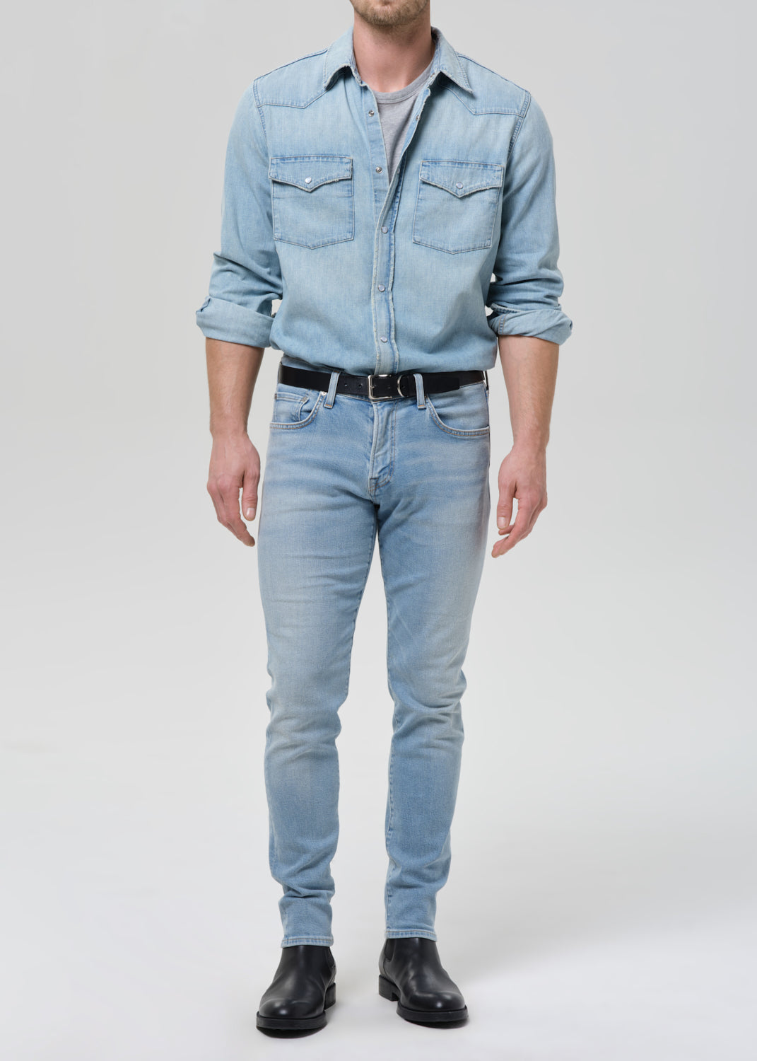 Man wearing light blue straight leg jeans with a light blue denim button-up shirt on a grey background.