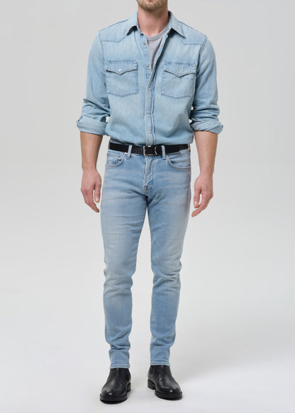 Man wearing light blue straight leg jeans with a light blue denim button-up shirt on a grey background.