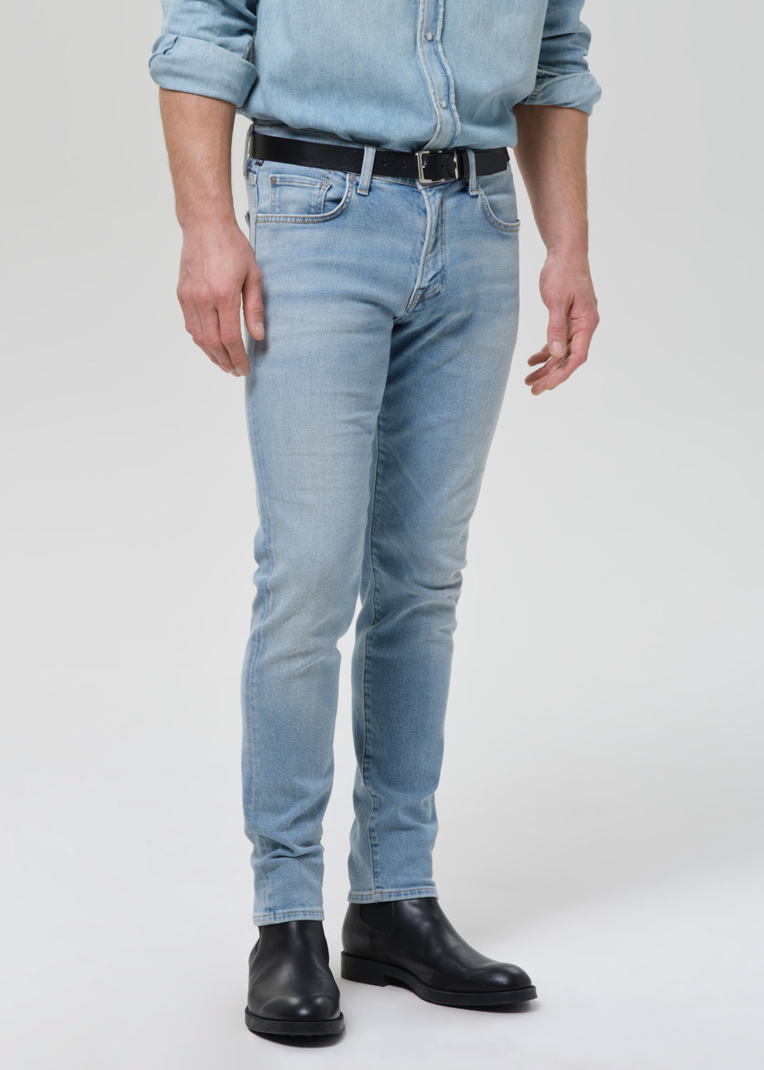 Man wearing light blue straight leg jeans with a light blue denim button-up shirt on a grey background.