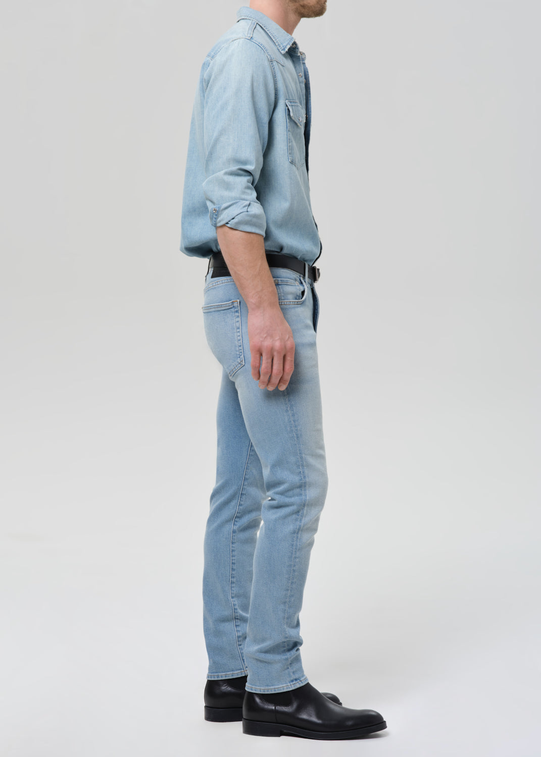 Man wearing light blue straight leg jeans with a light blue denim button-up shirt viewed from the side on a grey background.