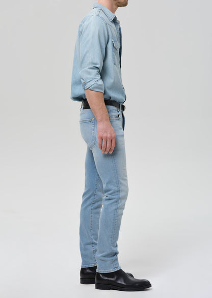 Man wearing light blue straight leg jeans with a light blue denim button-up shirt viewed from the side on a grey background.