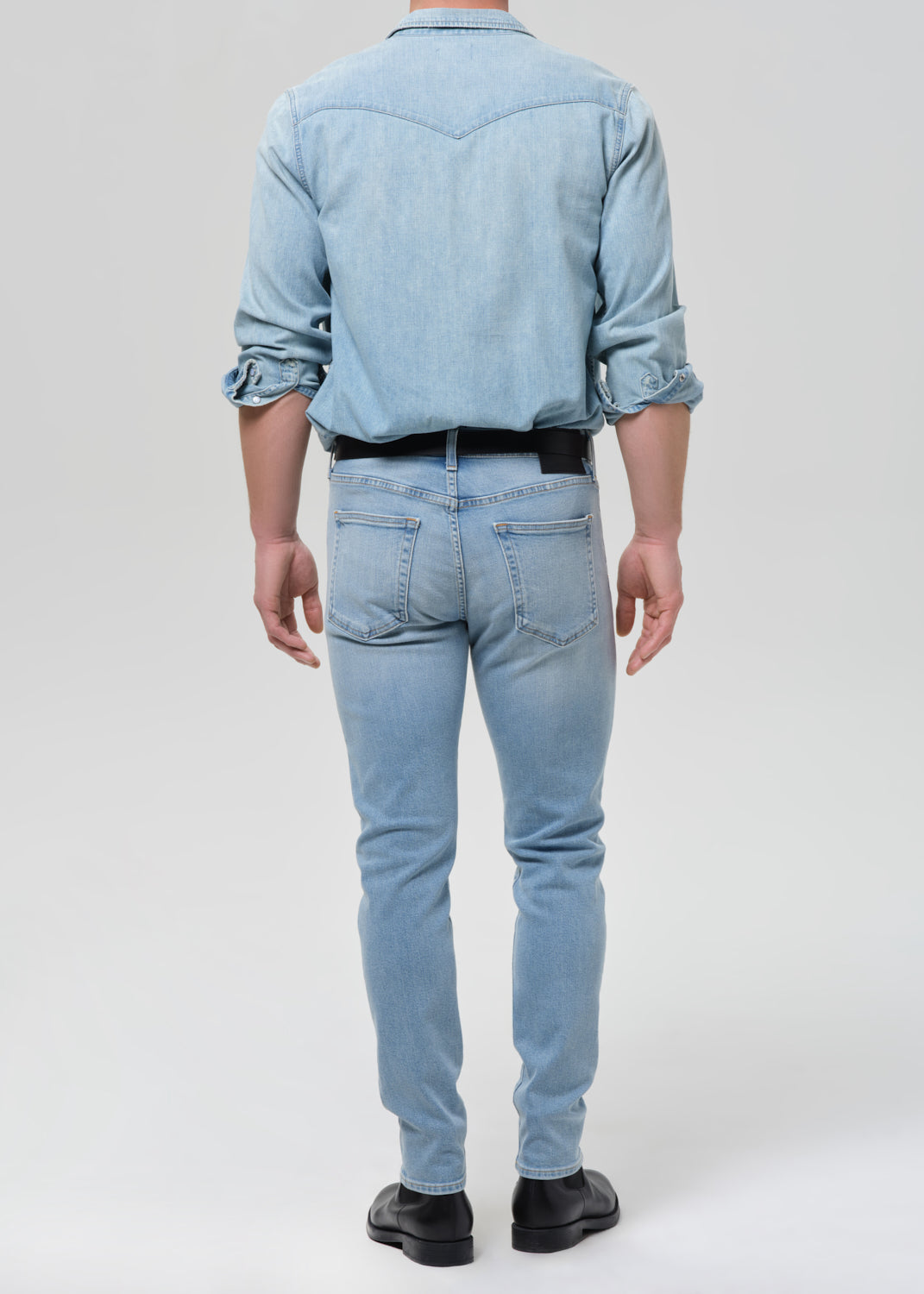 Man wearing light blue straight leg jeans with a light blue denim button-up shirt viewed from the back on a grey background.