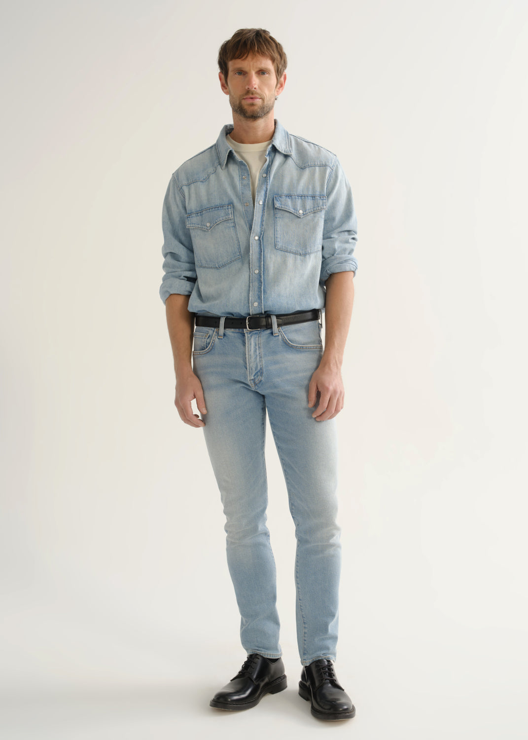 Person wearing light wash blue slim tapered jeans with a light blue denim shirt, white tee, black belt, and black shoes, front view.