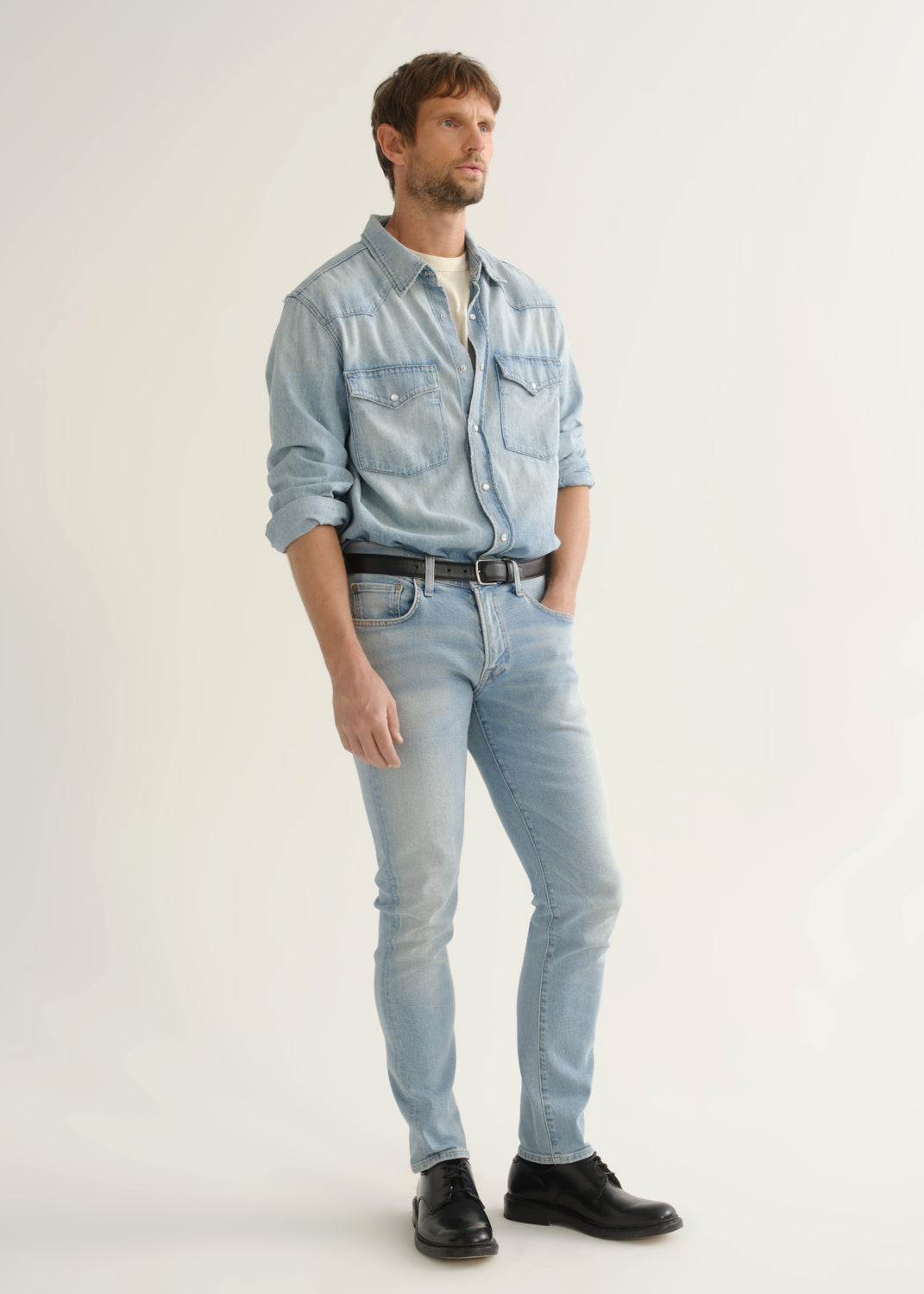 Person wearing light wash blue slim tapered jeans with a light blue denim shirt, white tee, black belt, and black shoes, side view.