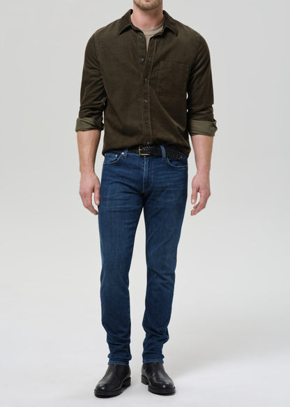 Man wearing medium blue straight leg jeans with an olive button-up shirt on a grey background.
