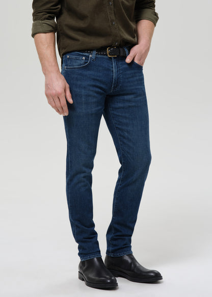 Man wearing medium blue straight leg jeans with an olive button-up shirt on a grey background.