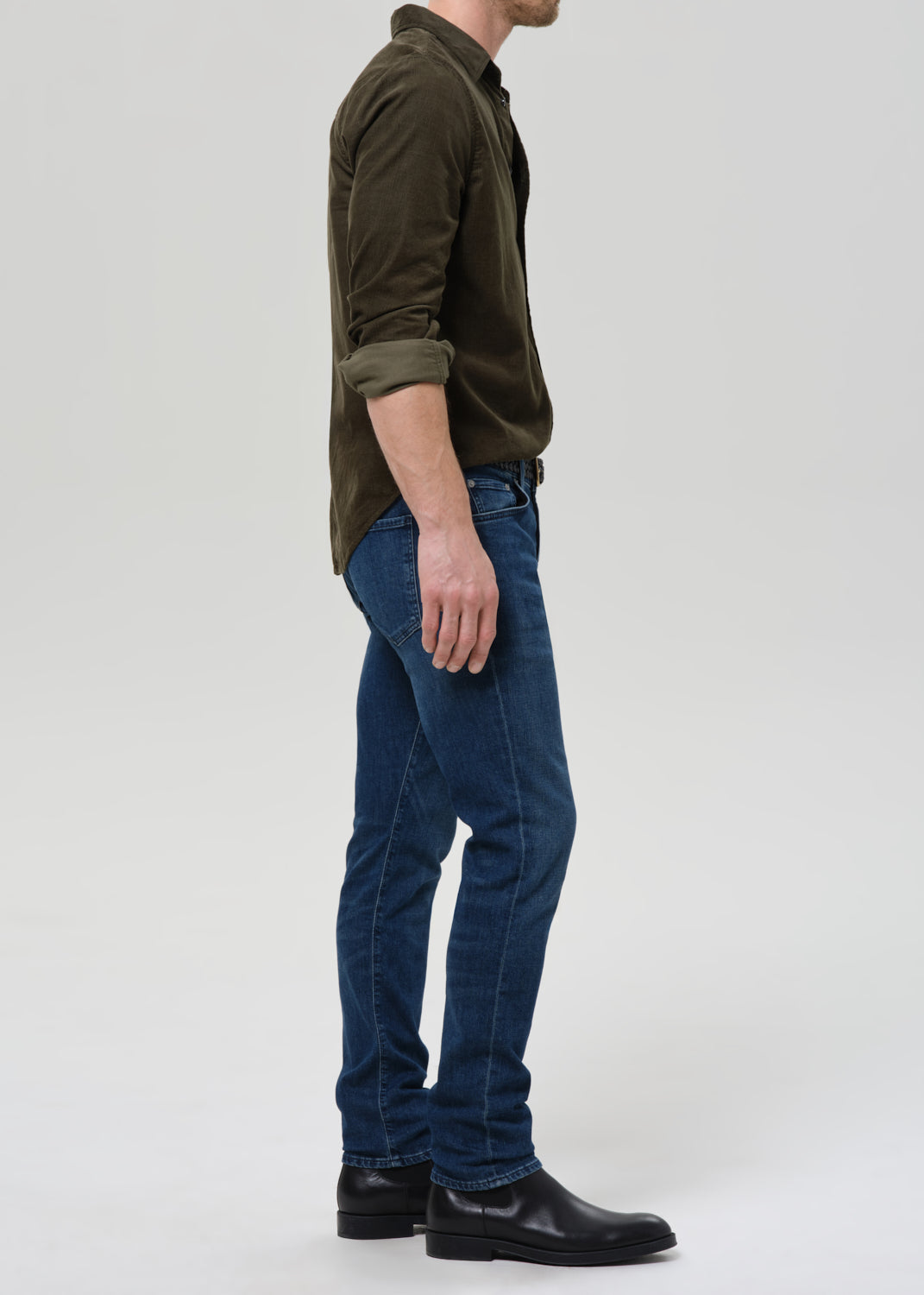 Man wearing medium blue straight leg jeans with an olive button-up shirt viewed from the side on a grey background.