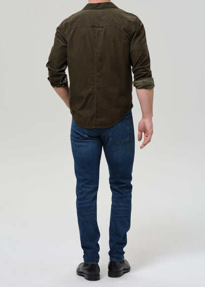 Man wearing medium blue straight leg jeans with an olive button-up shirt viewed from the back on a grey background.