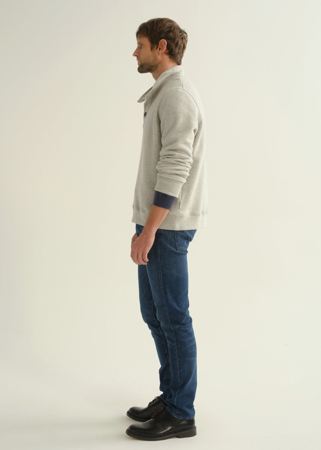 Person wearing dark blue slim tapered jeans with a light grey button-collar pullover, navy tee, and black shoes, back view