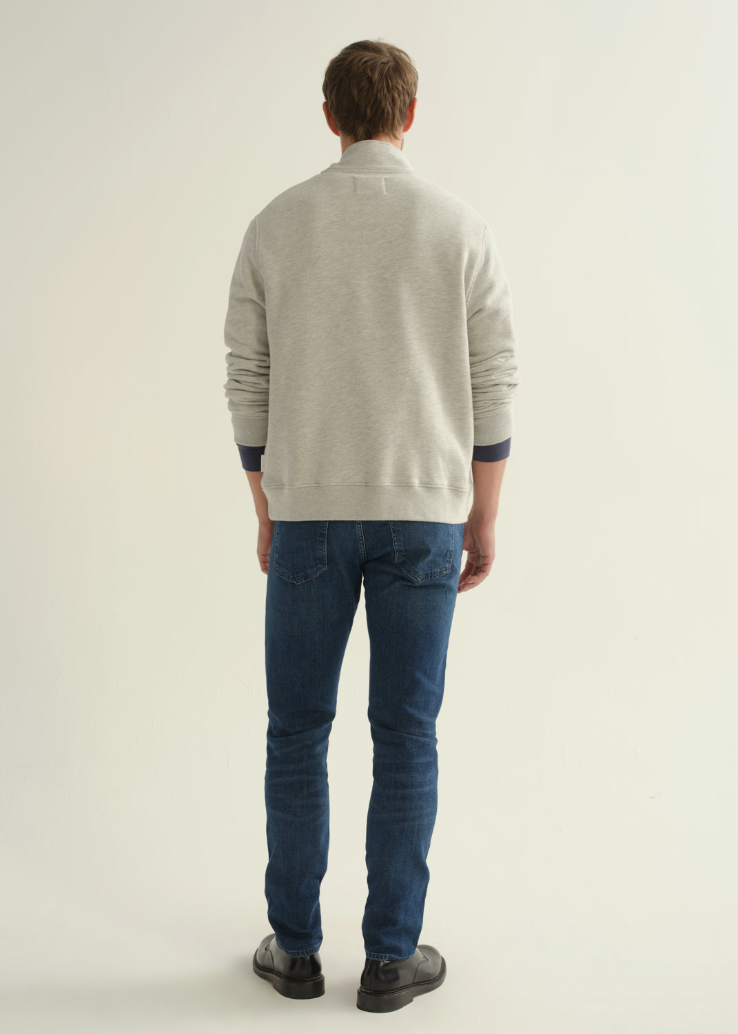Person wearing dark blue slim tapered jeans with a light grey button-collar pullover, navy tee, and black shoes, back view.