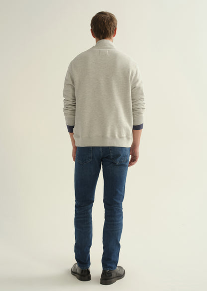 Person wearing dark blue slim tapered jeans with a light grey button-collar pullover, navy tee, and black shoes, back view.