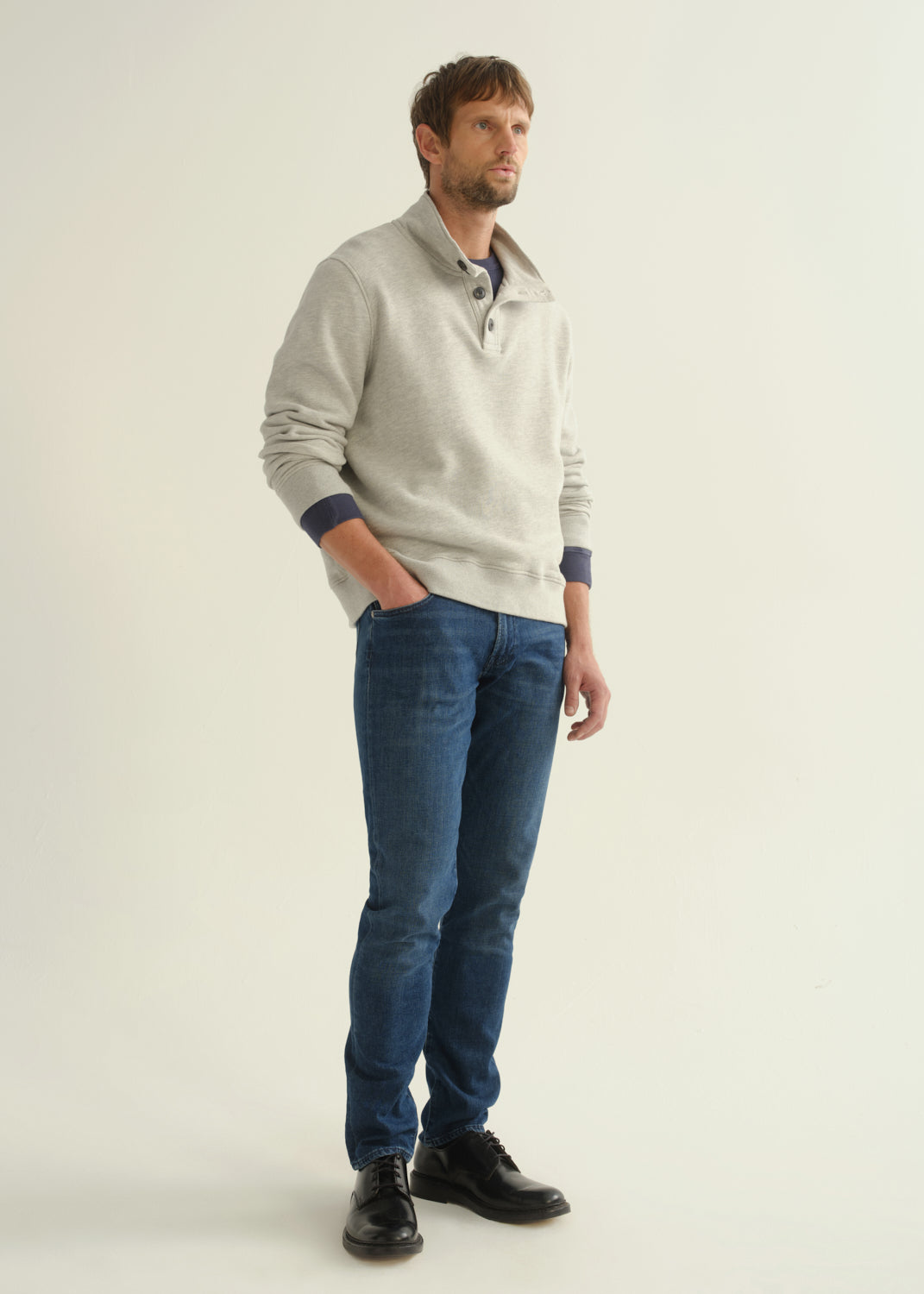 Person wearing dark blue slim tapered jeans with a light grey button-collar pullover, navy tee, and black shoes, side view.