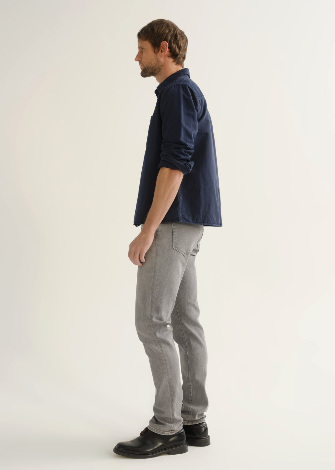 Person wearing light grey slim tapered jeans with a navy long sleeve button-up shirt and black shoes, side view.