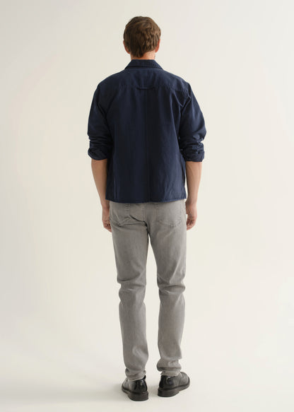 Person wearing light grey slim tapered jeans with a navy long sleeve button-up shirt layered over a white t-shirt and black shoes, front view.