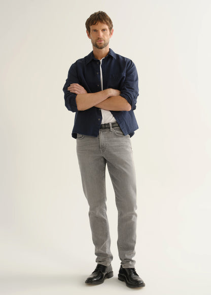 Person wearing light grey slim tapered jeans with a navy long sleeve button-up shirt layered over a white t-shirt and black shoes, front view.