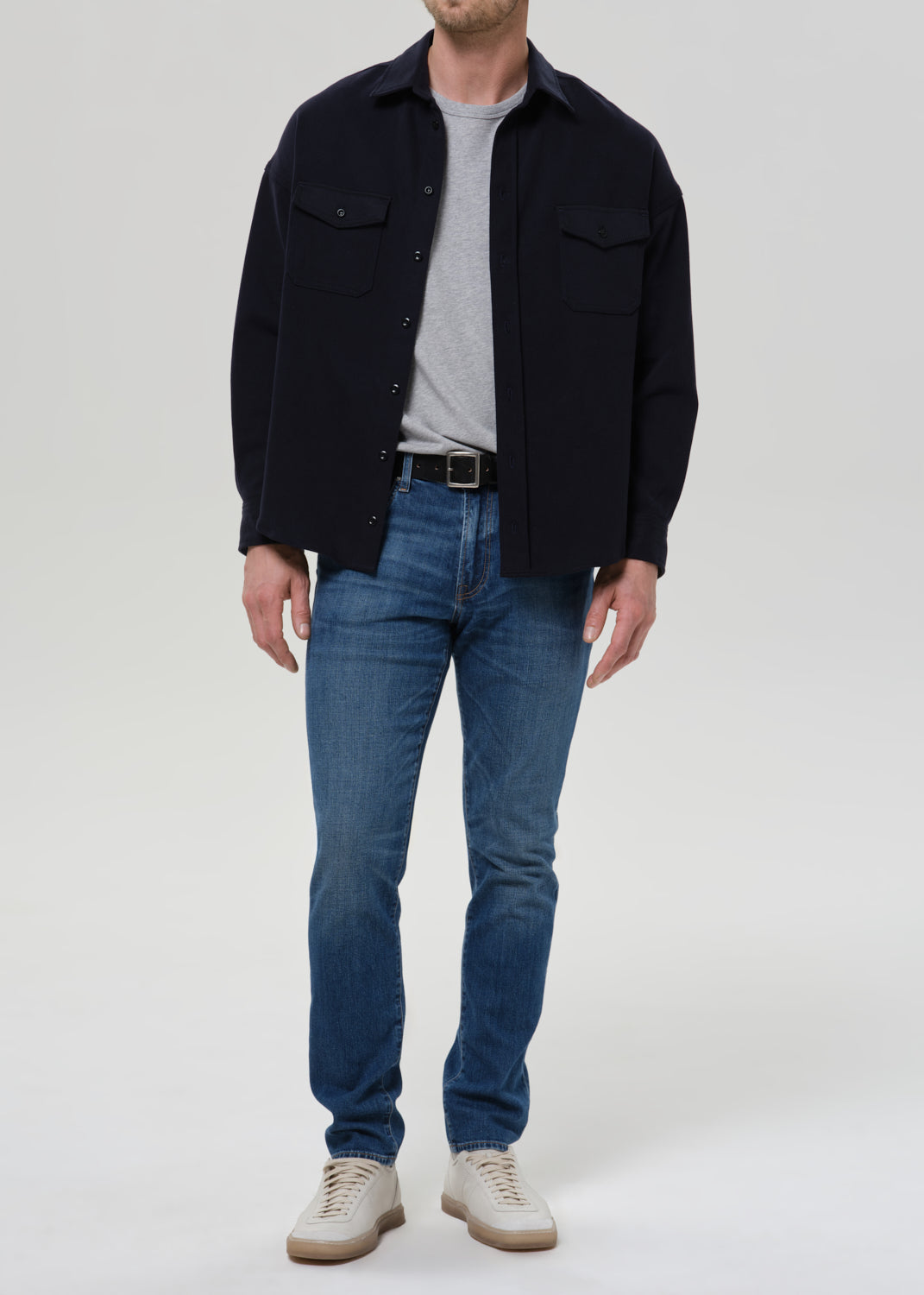 A man wearing a dark navy overshirt, blue jeans, a gray t-shirt, and off-white sneakers, standing against a grey background.