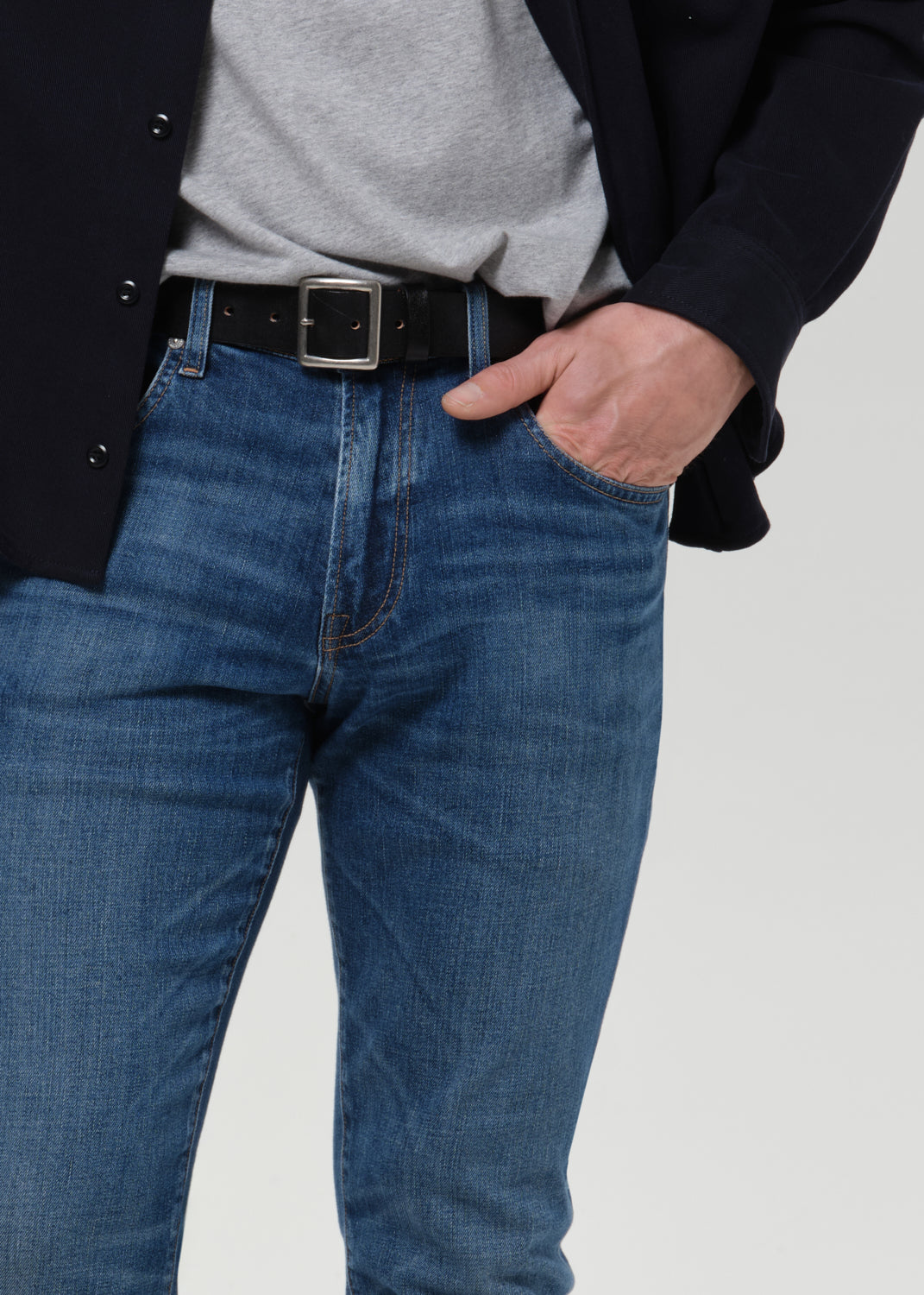 A close-up of a person wearing blue denim jeans with a black belt, a gray t-shirt, and a dark blazer, standing against a grey background.