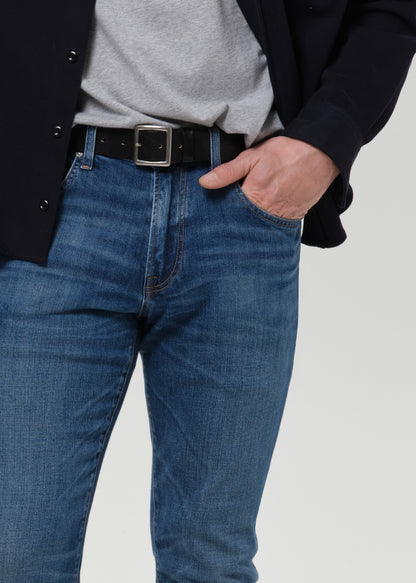 A close-up of a person wearing blue denim jeans with a black belt, a gray t-shirt, and a dark blazer, standing against a grey background.