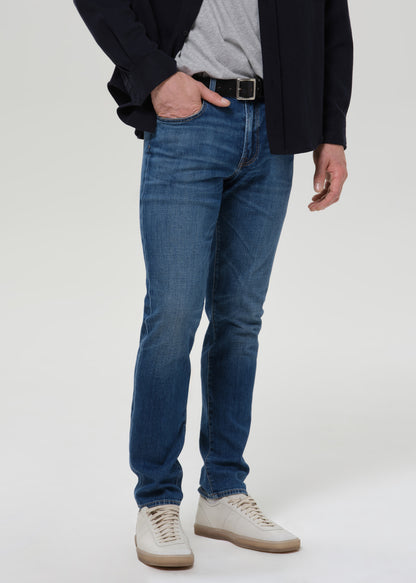 A person wearing slim-fit blue jeans with a black belt, a gray t-shirt, a black blazer, and off-white sneakers, shown from the waist down on a grey background.
