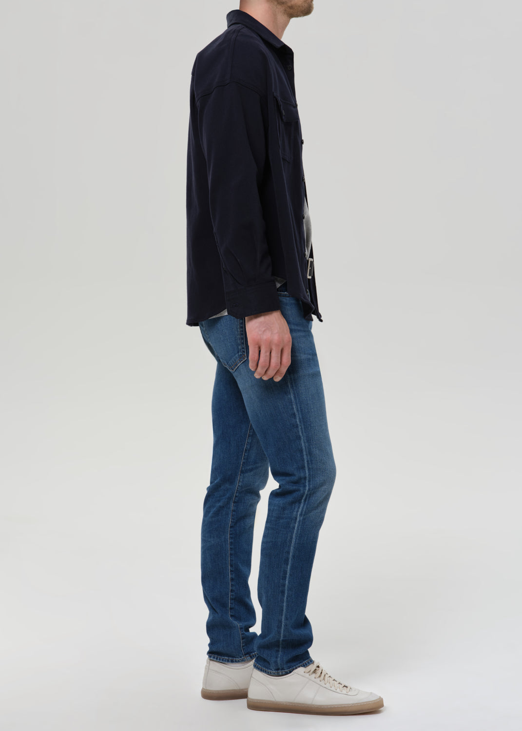A man shown in profile wearing blue slim-fit jeans, a dark navy overshirt, and off-white sneakers against a grey background.