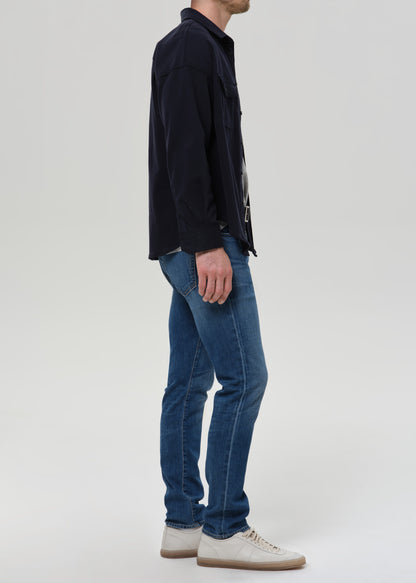 A man shown in profile wearing blue slim-fit jeans, a dark navy overshirt, and off-white sneakers against a grey background.