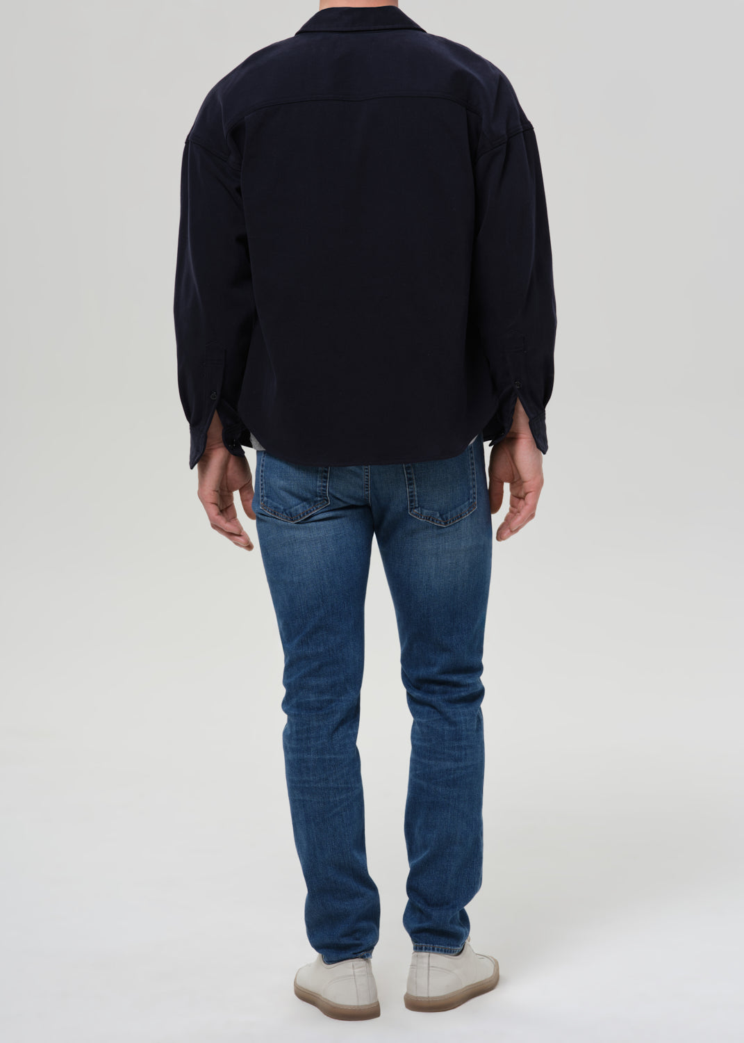 A man viewed from behind wearing a dark navy long-sleeve button-up shirt, blue jeans, and off-white slip-on shoes, standing against a grey background.