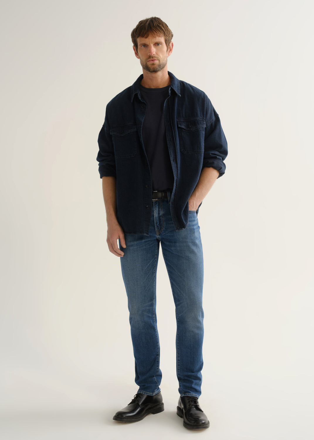 Person wearing a dark navy denim overshirt layered over a navy crewneck tee, medium blue slim tapered jeans, and black leather shoes, front full body view on a light grey background.