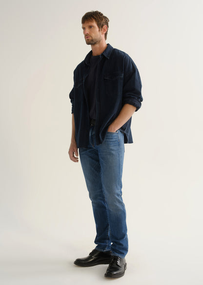 Person wearing a dark navy denim overshirt with rolled sleeves, medium blue slim tapered jeans, and black leather shoes, side profile full body view on a light grey background.