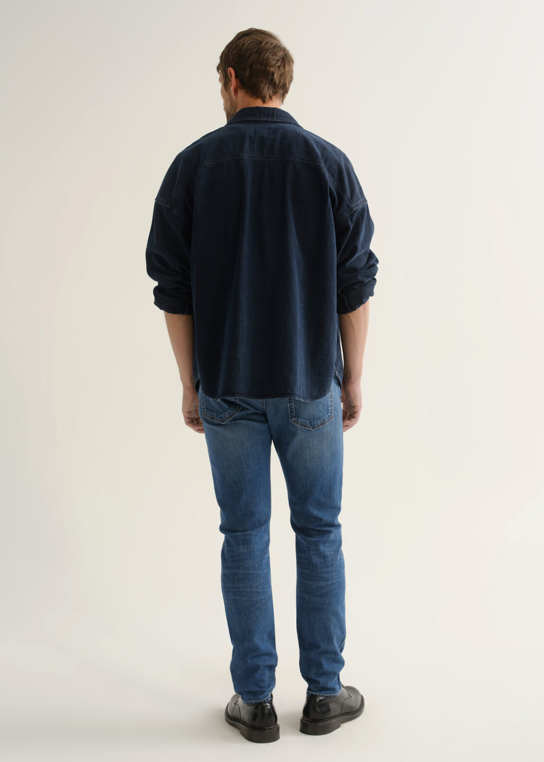 Person wearing a dark navy denim overshirt, medium blue slim tapered jeans, and black leather shoes, back full body view on a light grey background.