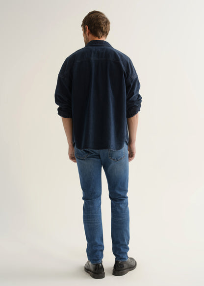 Person wearing a dark navy denim overshirt, medium blue slim tapered jeans, and black leather shoes, back full body view on a light grey background.